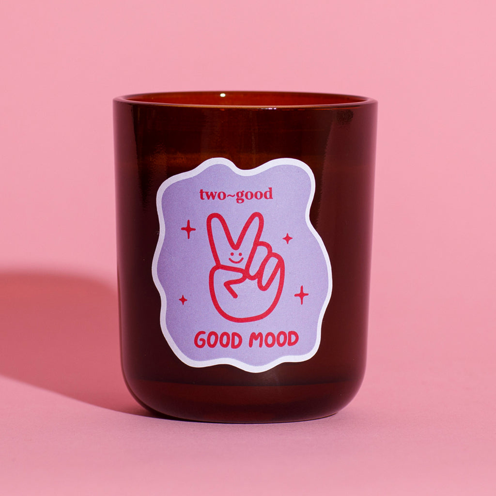 Brown glass candle vessel with a 'two-good GOOD MOOD' label on a pink background illustrations are linear and bold