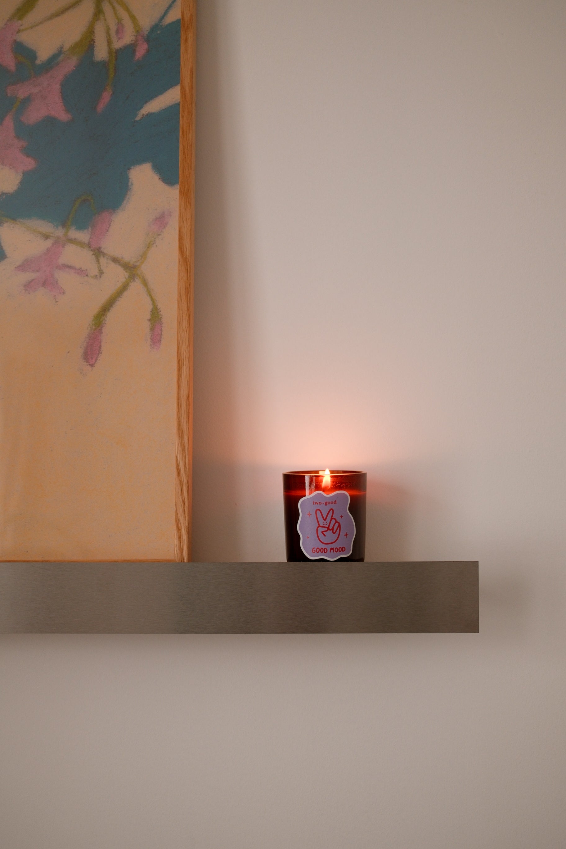 A lit amber glass soy wax candle with a purple peace sign sticker sits on a dark shelf against a beige wall, next to a framed floral artwork.
