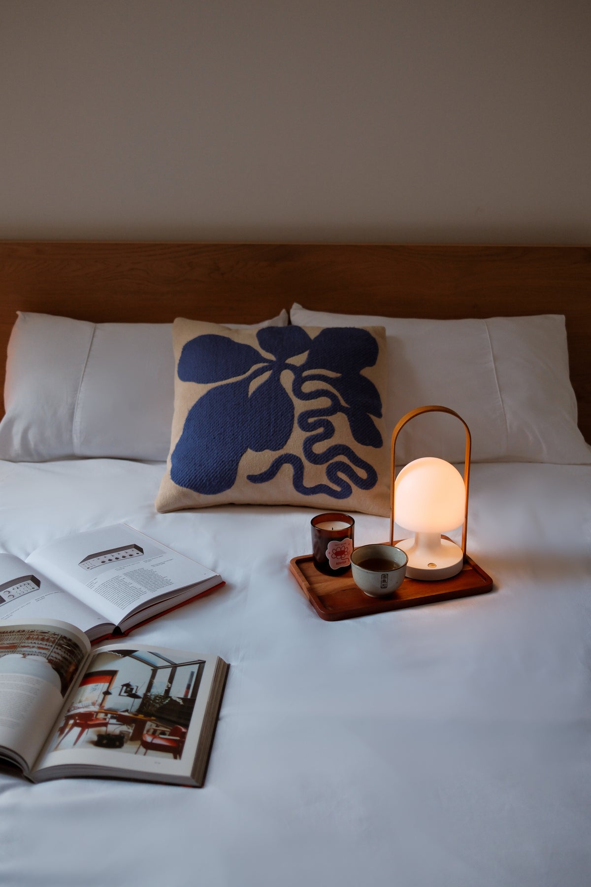 A styled bedroom scene with a bed made of white linens, a wooden headboard, a cream pillow with a bold blue abstract print, an open book, a small tray with a glowing warm lamp and cup, and a second open book in the foreground.