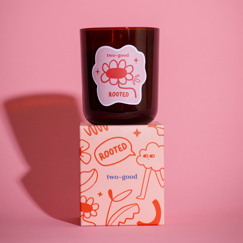 Brown glass candle vessel with 'two-good' branding on a pink background illustrations on packaging fun soy wax candle