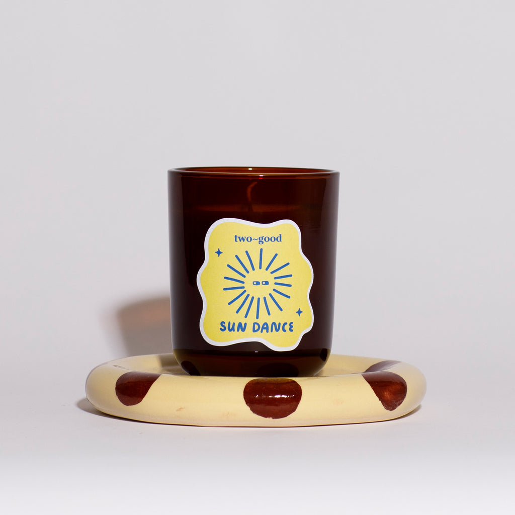 Brown glass candle vessel with illustrations on label on a polka dot stand against a light grey background 