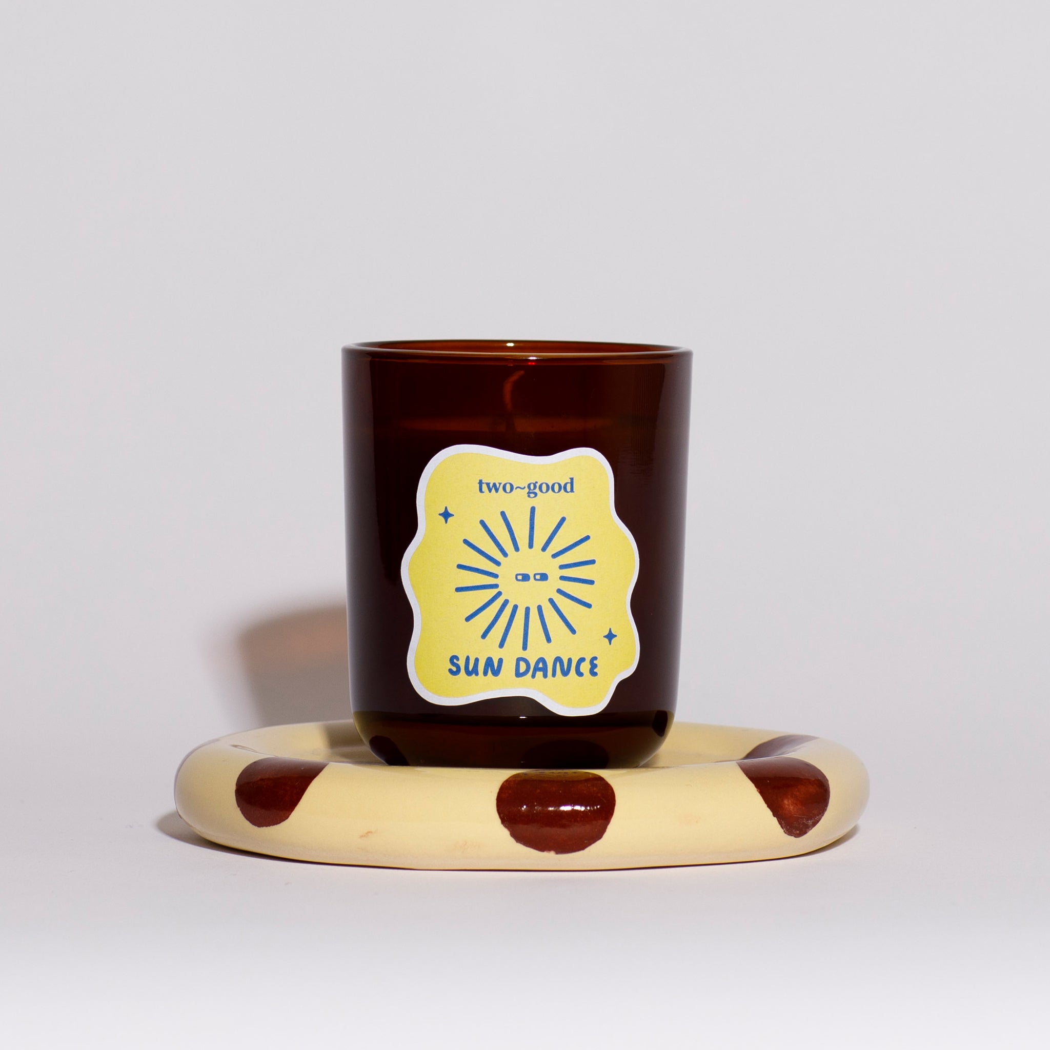 Brown glass candle vessel with illustrations on label on a polka dot stand against a light grey background 