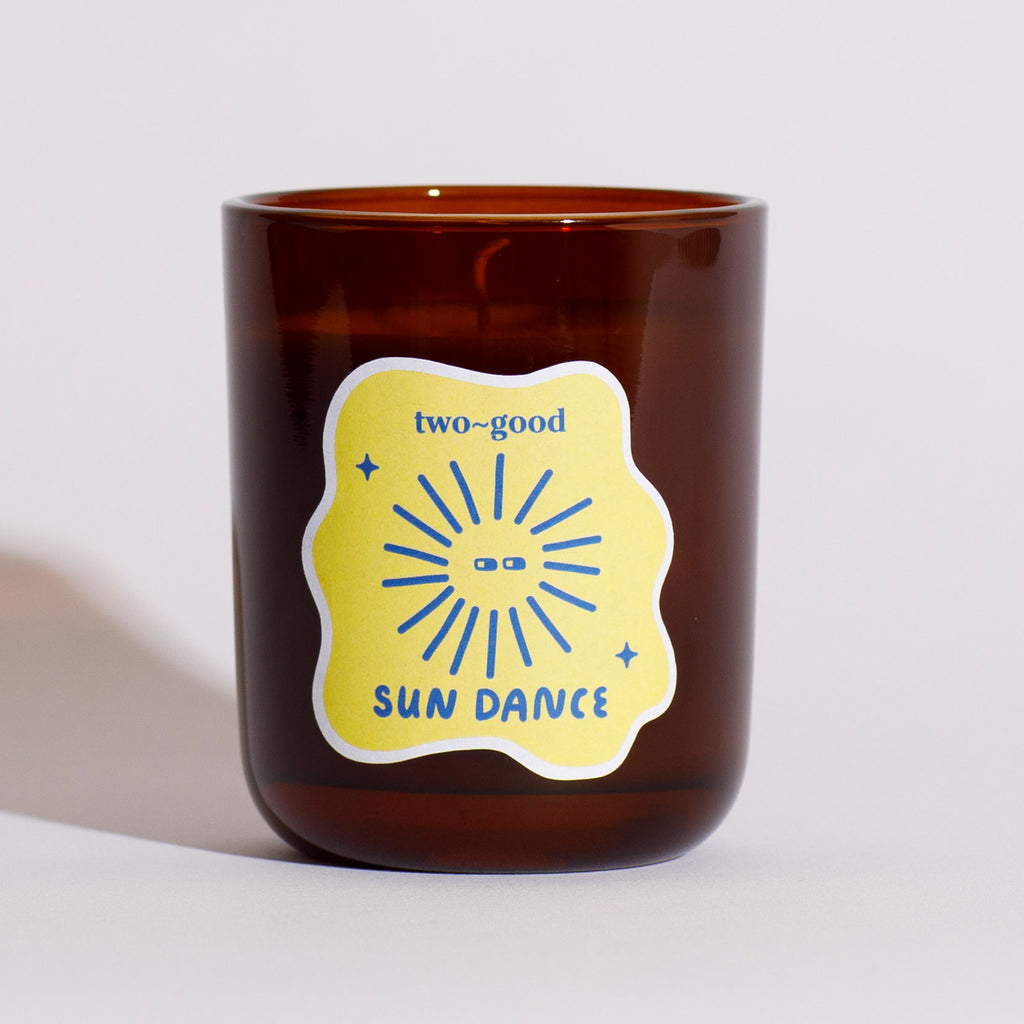 Brown glass candle vessel with a yellow label featuring 'two-good' and 'Sun Dance' text on a light grey background. Soy wax candle. Tropical scent, fruity coconut