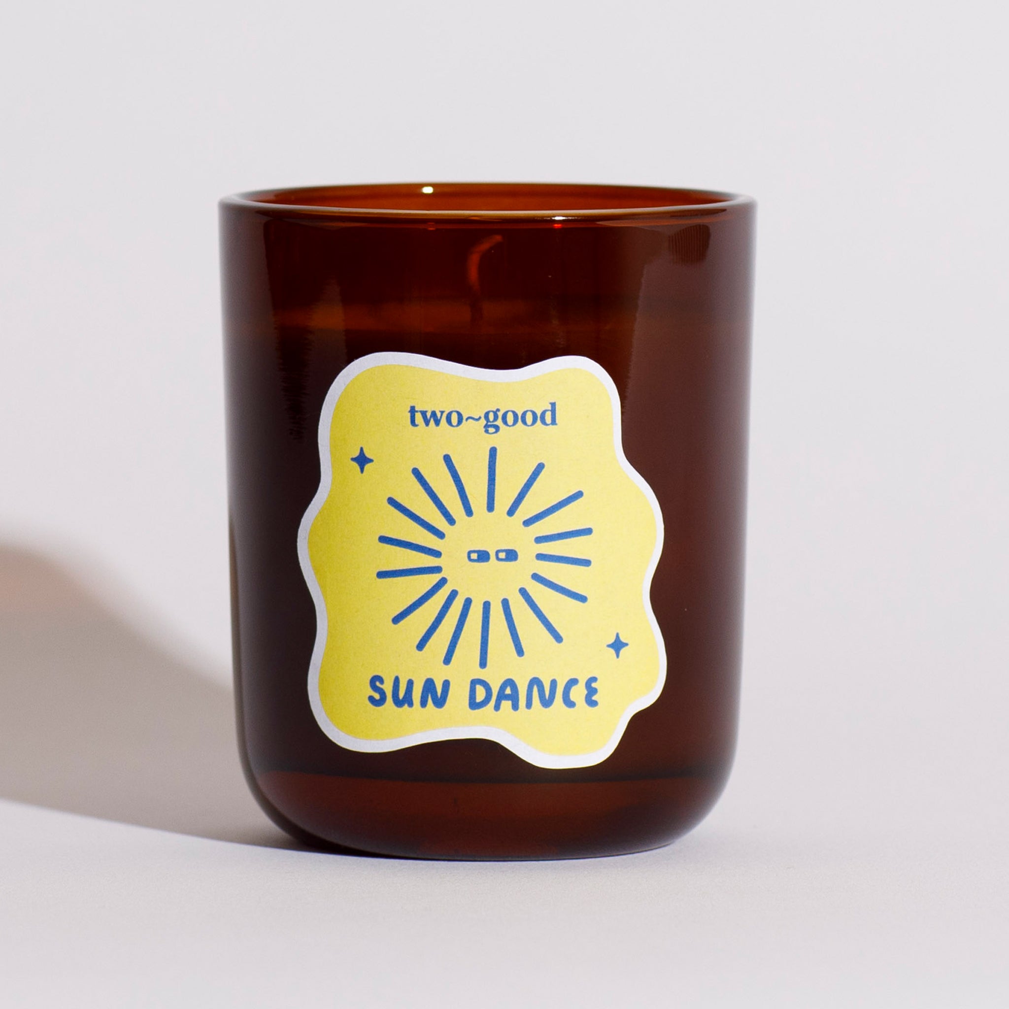 Brown glass candle vessel with a yellow label featuring 'two-good' and 'Sun Dance' text on a light grey background. Soy wax candle. Tropical scent, fruity coconut