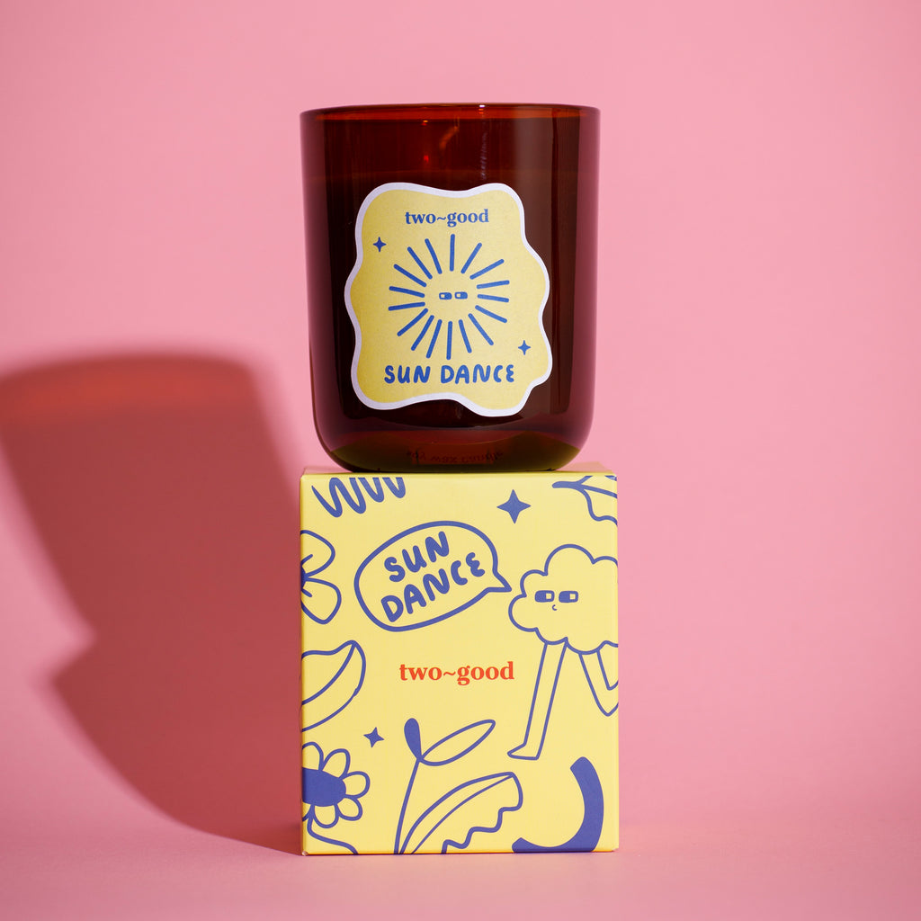 Brown glass candle vessel with 'two-good' branding on a pink background. Box packaging with linear illustrations. 