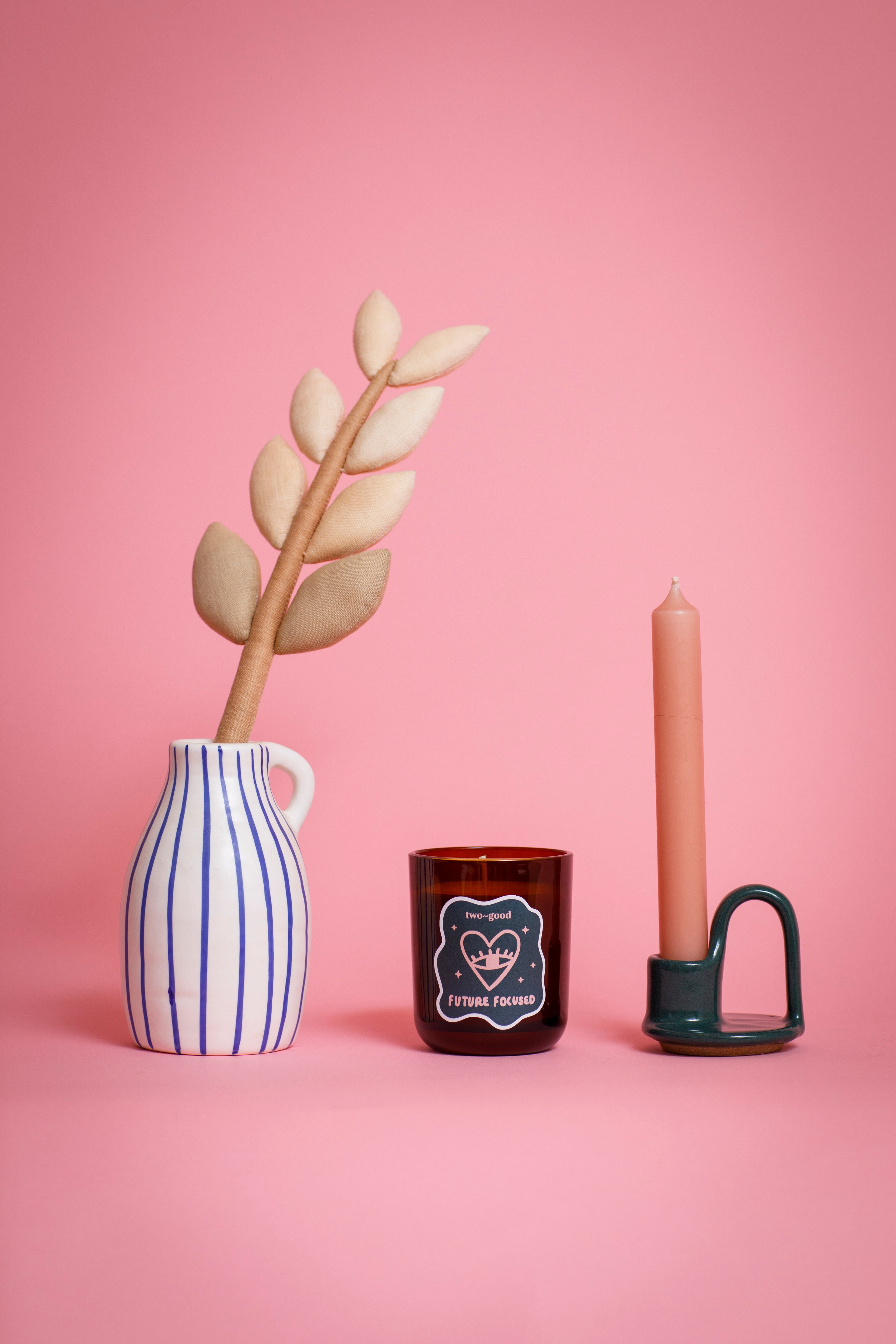 Decorative items including a striped vase with a plant, a candle in a holder, and a small glass soy wax candle in jar on a pink background.
