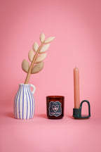 Decorative items including a striped vase with a plant, a candle in a holder, and a small glass soy wax candle in jar on a pink background.