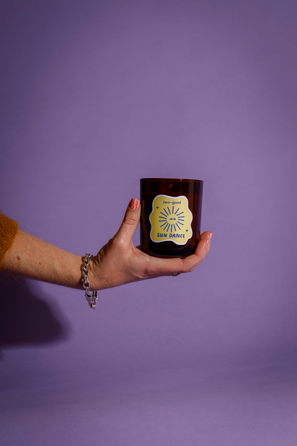 Hand holding a dark glass candle vessel with a illustrations on label against a purple background