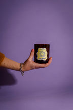 Hand holding a dark glass candle vessel with a illustrations on label against a purple background