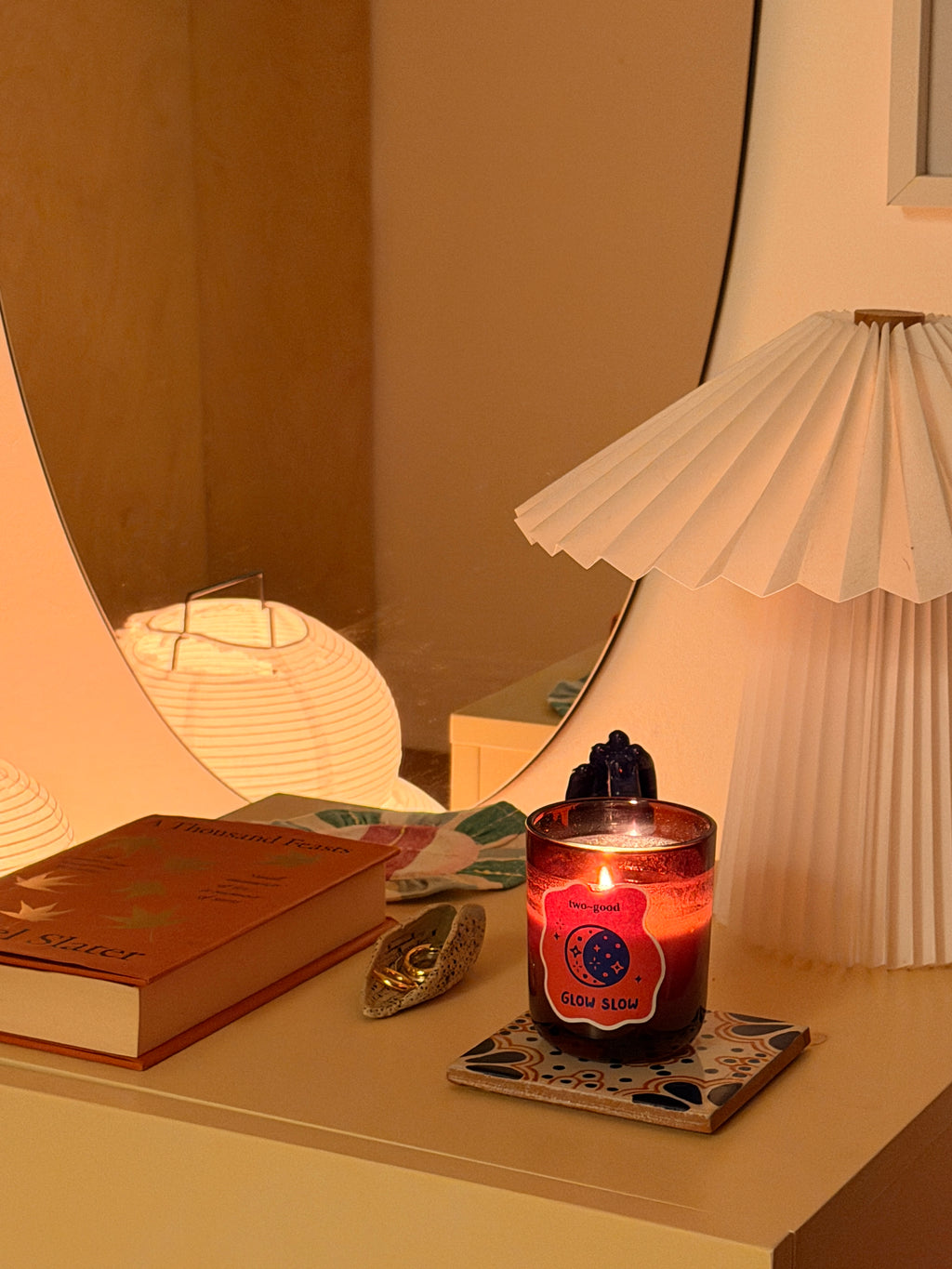 A lit candle in a dark glass jar sits on a tabletop beside a folded orange book, a small metallic object, and a pleated white table lamp in a warm indoor setting.